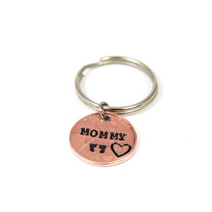 Puede incluir: Un llavero de cobre con las palabras "Mommy" y dos huellas de pies, y un coraz&oacute;n grabado en &eacute;l.