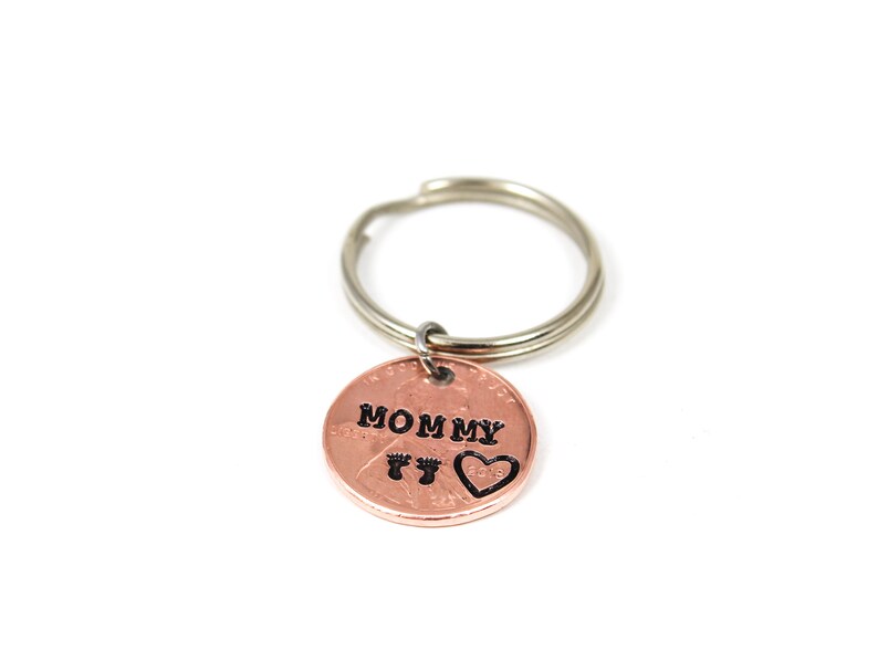 Puede incluir: Un llavero de cobre con las palabras "Mommy" y dos huellas de pies, y un coraz&oacute;n grabado en &eacute;l.