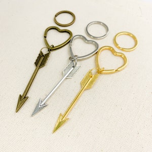 Op de afbeelding: Drie gouden sleutelhangers met pijl-bedels. De sleutelhangers zijn hartvormig. De pijl-bedels hebben een eenvoudig ontwerp met een puntige punt en een klein veerdetail.
