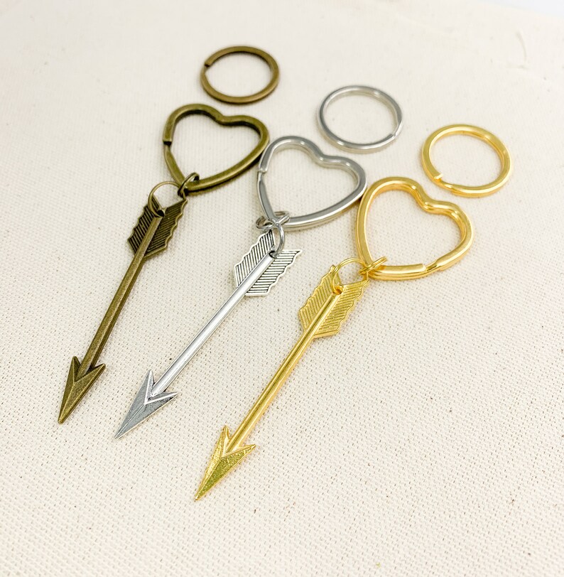 Op de afbeelding: Drie gouden sleutelhangers met pijl-bedels. De sleutelhangers zijn hartvormig. De pijl-bedels hebben een eenvoudig ontwerp met een puntige punt en een klein veerdetail.