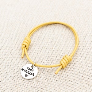 May include: A yellow leather bracelet with a silver charm that reads "endo warrior" with a small heart.