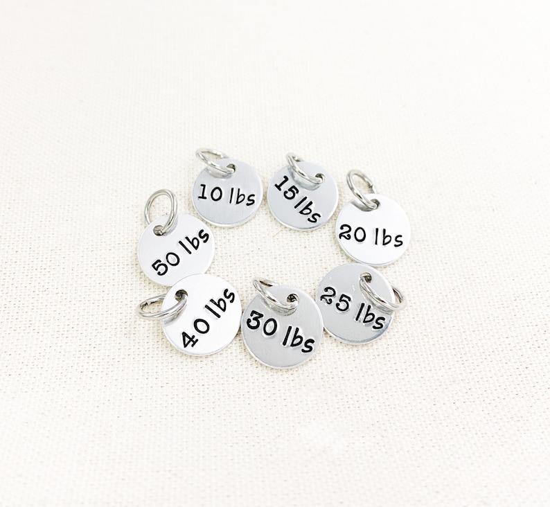 May include: Set of seven silver metal weight tags with black lettering. The tags are in a circle and show the following weights: 10 lbs, 15 lbs, 20 lbs, 25 lbs, 30 lbs, 40 lbs, and 50 lbs.