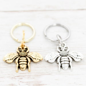 Puede incluir: Dos llaveros con forma de abeja, uno dorado y otro plateado, con un dise&ntilde;o de anillo simple.
