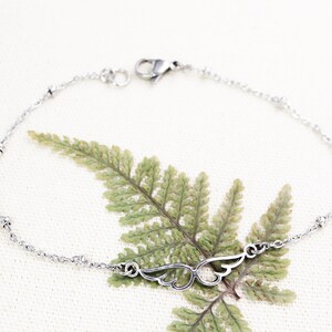 May include: A silver chain bracelet with small beads and a delicate wing charm. The bracelet is on a white background with a green fern leaf.
