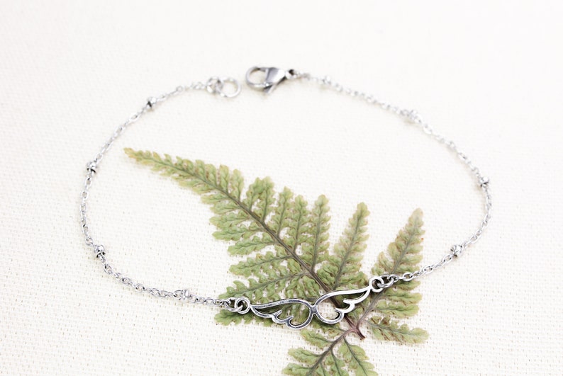 May include: A silver chain bracelet with small beads and a delicate wing charm. The bracelet is on a white background with a green fern leaf.