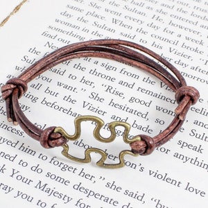 May include: A brown leather bracelet with a metal puzzle piece charm. The bracelet is tied with a knot and has a simple design.