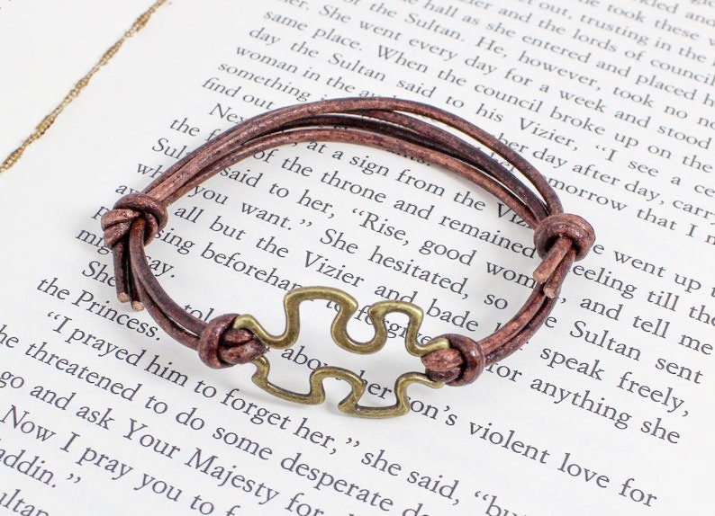 May include: A brown leather bracelet with a metal puzzle piece charm. The bracelet is tied with a knot and has a simple design.