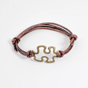 May include: A brown leather bracelet with a gold-toned puzzle piece charm.