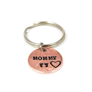 Puede incluir: Llavero de un centavo de cobre con un llavero de plata. El centavo tiene las palabras "Mommy" y un coraz&oacute;n con huellas de pies grabadas.