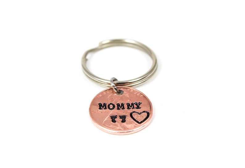 Puede incluir: Llavero de un centavo de cobre con un llavero de plata. El centavo tiene las palabras "Mommy" y un coraz&oacute;n con huellas de pies grabadas.