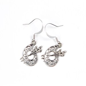 May include: Silver earrings shaped like paint palettes with a paintbrush on each. The earrings have a hook closure.