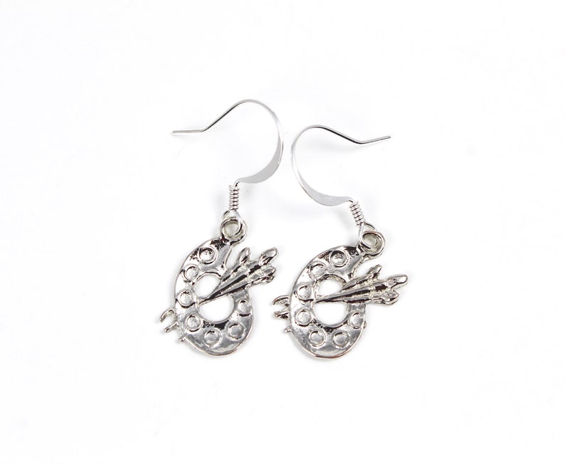 May include: Silver earrings shaped like paint palettes with a paintbrush on each. The earrings have a hook closure.