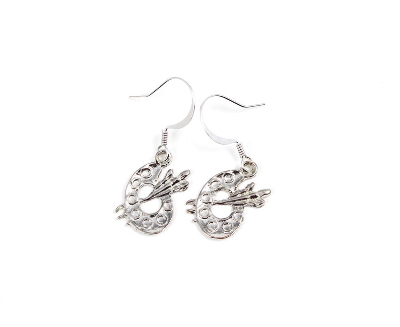 May include: Silver earrings shaped like an artist's palette with a paintbrush on top.