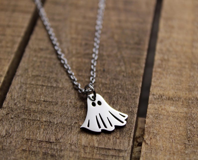 May include: A silver-colored necklace with a ghost pendant. The ghost pendant has two eye holes and a wavy bottom edge. The necklace is on a wooden surface.