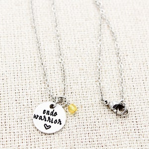 May include: Silver necklace with a round pendant that says "endo warrior" with a small heart. A small yellow bead hangs from the pendant.