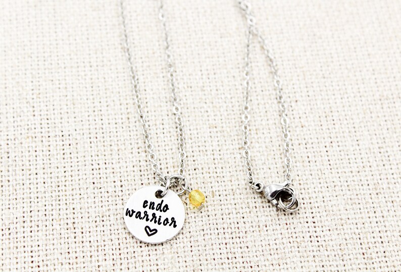 May include: Silver necklace with a round pendant that says "endo warrior" with a small heart. A small yellow bead hangs from the pendant.