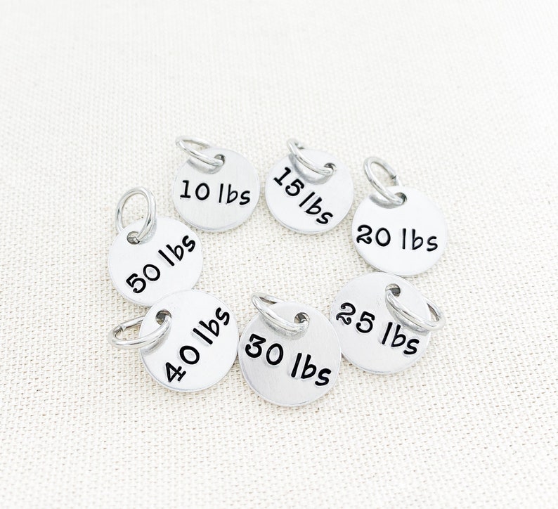 May include: Set of seven silver metal weight tags with black lettering. The tags are labeled with the following weights: 10 lbs, 15 lbs, 20 lbs, 25 lbs, 30 lbs, 40 lbs, and 50 lbs.