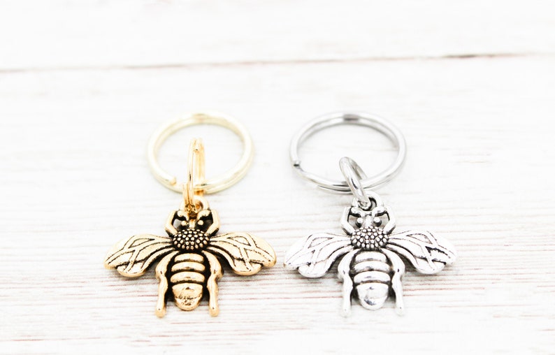 Puede incluir: Dos llaveros con forma de abeja, uno dorado y otro plateado, con un dise&ntilde;o detallado. Los llaveros est&aacute;n unidos a llaveros de plata.