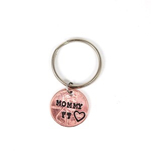 Puede incluir: Un llavero de un centavo de cobre con la palabra "MOMMY" y dos huellas de pies grabadas. Un peque&ntilde;o coraz&oacute;n est&aacute; grabado al lado de las huellas.