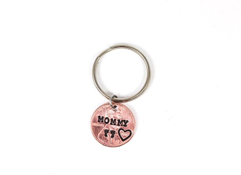 Puede incluir: Un llavero de un centavo de cobre con la palabra "MOMMY" y dos huellas de pies grabadas. Un peque&ntilde;o coraz&oacute;n est&aacute; grabado al lado de las huellas.