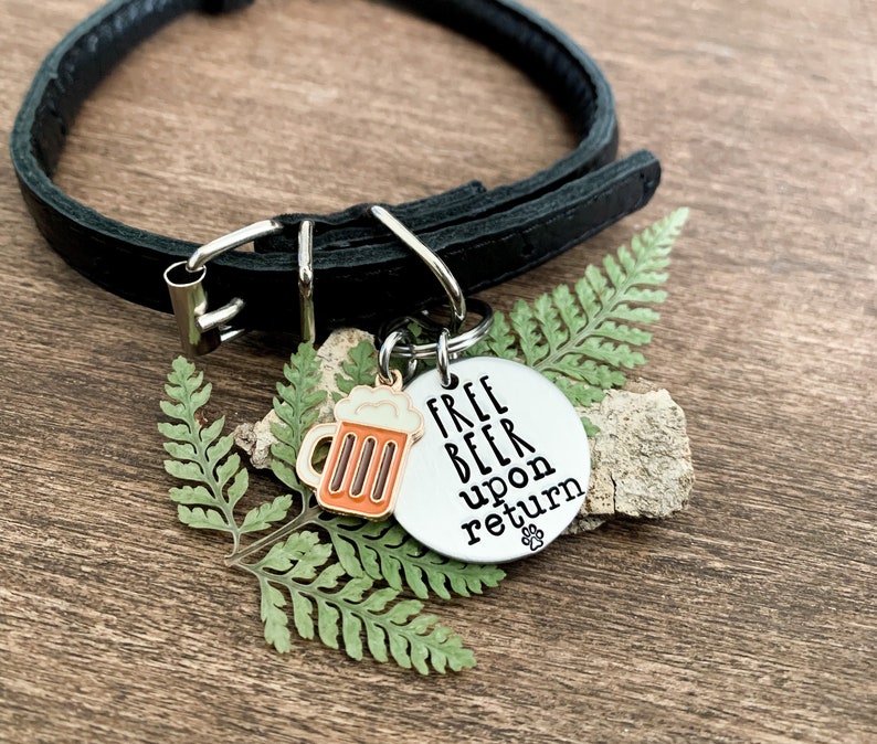 K&ouml;nnte beinhalten: Ein silbernes Hundehalsband mit dem Text "Free Beer Upon Return" und einem kleinen Bierkrug-Symbol. Das Schild ist an einem schwarzen Lederhundekragen befestigt.