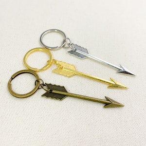 Op de afbeelding: Drie pijlvormige sleutelhangers met verschillende metalen afwerkingen. De bovenste sleutelhanger is zilver, de middelste is goud en de onderste is brons.
