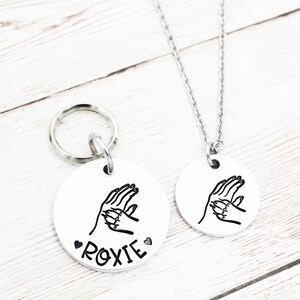 May include: A silver dog tag with a paw print design and the name "ROXIE" engraved on it. The tag is attached to a silver key ring. A matching silver necklace with the same paw print design is also shown.