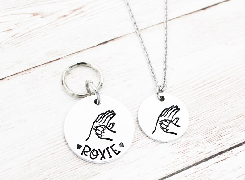 May include: A silver dog tag with a paw print design and the name "ROXIE" engraved on it. The tag is attached to a silver key ring. A matching silver necklace with the same paw print design is also shown.
