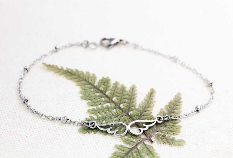 May include: A silver chain bracelet with a delicate wing charm. The bracelet has small beads along the chain.