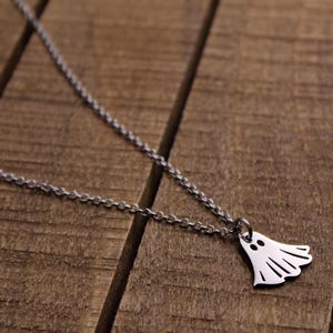 May include: A silver-colored necklace with a ghost pendant. The ghost pendant has two eye holes and a wavy bottom edge. The necklace chain is a delicate link style, and the background is a wooden surface.