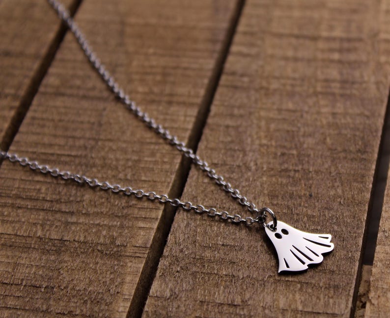 May include: A silver-colored necklace with a ghost pendant. The ghost pendant has two eye holes and a wavy bottom edge. The necklace chain is a delicate link style, and the background is a wooden surface.