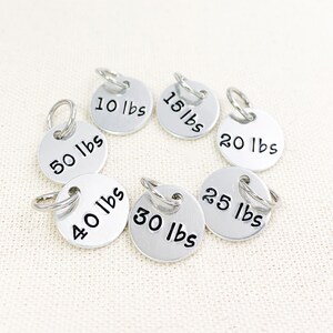May include: Set of seven silver metal weight tags with key rings. The tags are labeled with the following weights: 10 lbs, 15 lbs, 20 lbs, 25 lbs, 30 lbs, 40 lbs, and 50 lbs.