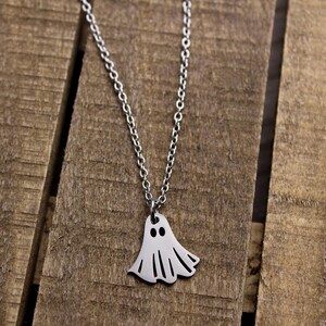 May include: A silver-colored necklace with a ghost pendant. The ghost pendant has two black eyes and a flowing, sheet-like body. The necklace chain is delicate and hangs against a wooden background.