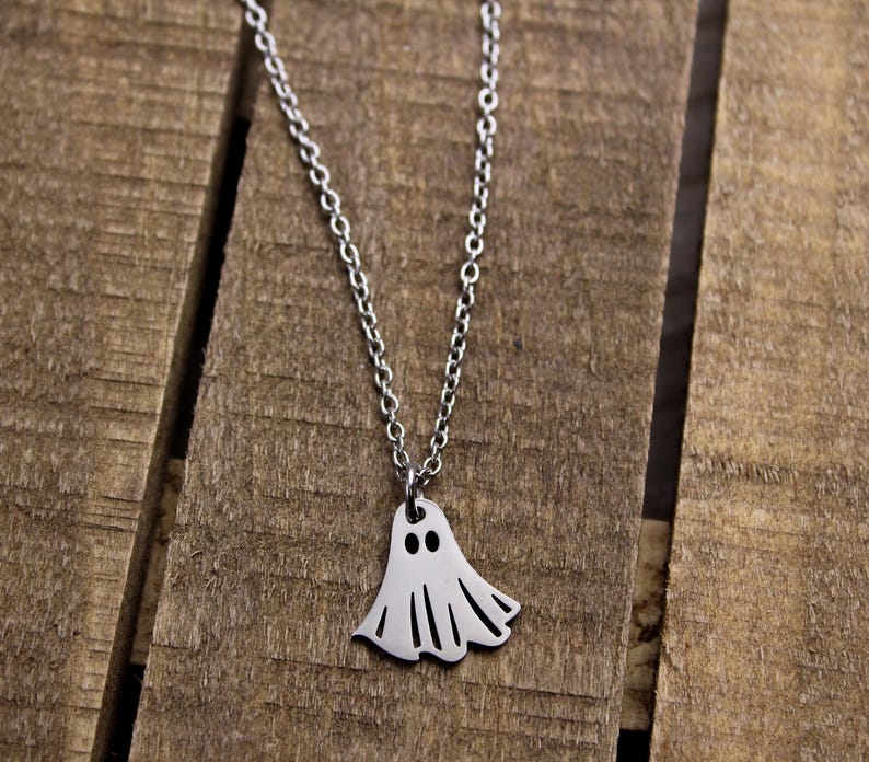 May include: A silver-colored necklace with a ghost pendant. The ghost pendant has two black eyes and a flowing, sheet-like body. The necklace chain is delicate and hangs against a wooden background.