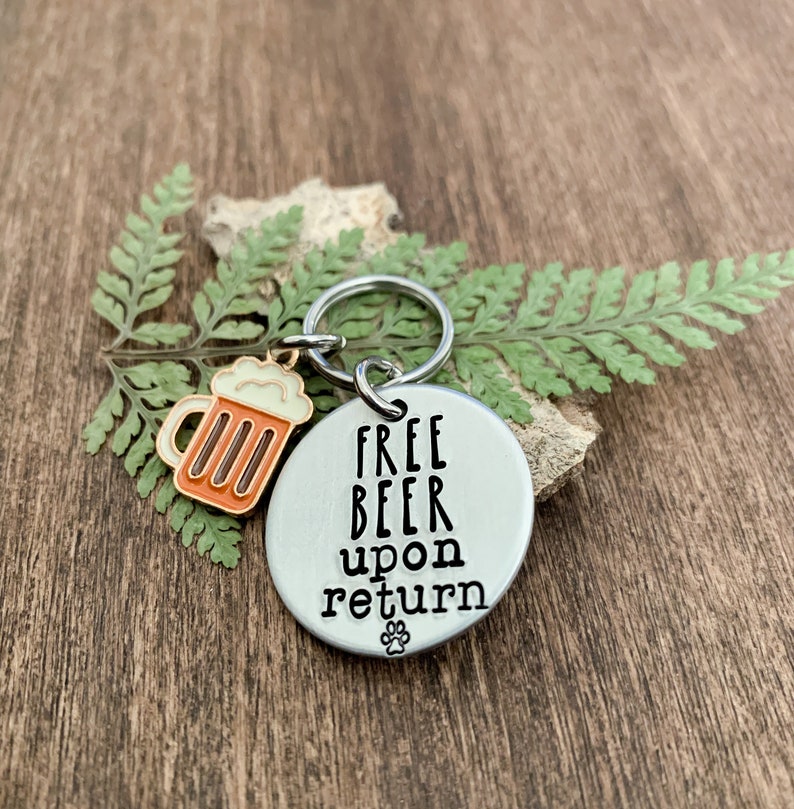 K&ouml;nnte beinhalten: Ein silberner Schl&uuml;sselanh&auml;nger mit einem Bierkrug-Anh&auml;nger und dem Text "FREE BEER upon return" mit einem Pfotenabdruck unter dem Text.