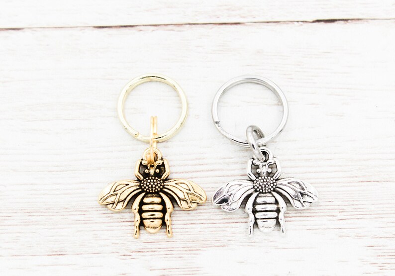 Puede incluir: Dos llaveros con forma de abeja, uno dorado y otro plateado, con un simple anillo de llavero unido a cada uno.