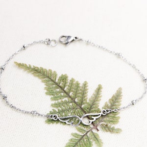 May include: A silver chain bracelet with a delicate wing charm. The chain has small round beads and a lobster clasp closure.