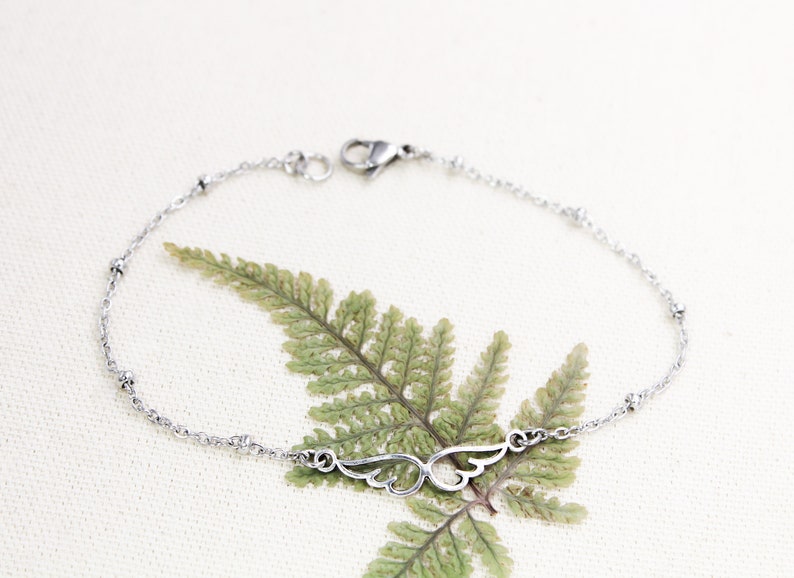 May include: A silver chain bracelet with a delicate wing charm. The chain has small round beads and a lobster clasp closure.