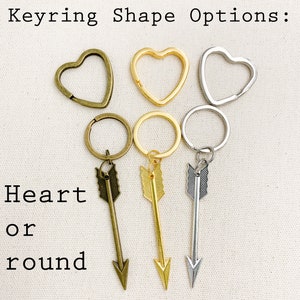 Op de afbeelding: Drie sleutelhangers met pijlbedels. De sleutelhangers zijn verkrijgbaar in hart- of ronde vormen. De hartvormen zijn in goud, zilver en brons. De ronde vormen zijn in goud, zilver en brons.