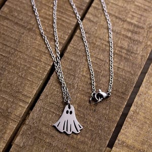May include: A silver-colored necklace with a ghost pendant. The ghost pendant has two eye holes and a wavy bottom edge. The necklace is on a wooden surface.