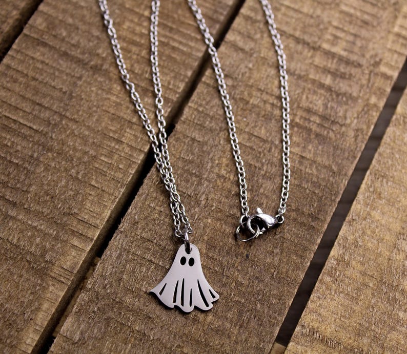 May include: A silver-colored necklace with a ghost pendant. The ghost pendant has two eye holes and a wavy bottom edge. The necklace is on a wooden surface.