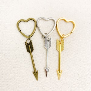 Op de afbeelding: Drie hartvormige sleutelhangers met pijl-bedels. De sleutelhangers zijn zilver, goud en brons.
