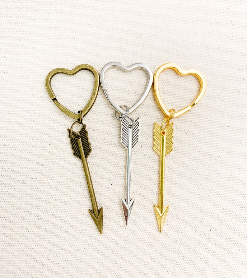 Op de afbeelding: Drie hartvormige sleutelhangers met pijl-bedels. De sleutelhangers zijn zilver, goud en brons.