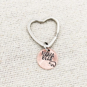 May include: A silver heart-shaped keychain with a copper penny attached. The penny has the words "Mama Bear" engraved on it, along with a small bear silhouette.