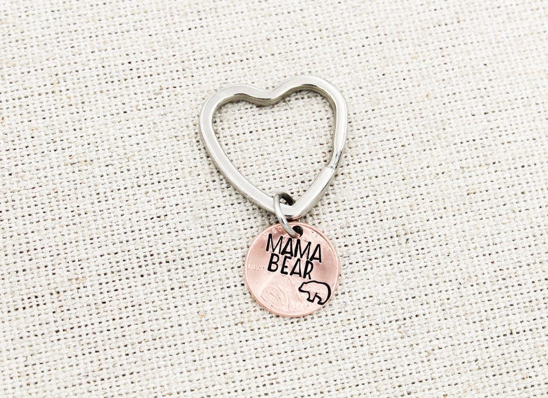 May include: A silver heart-shaped keychain with a copper penny attached. The penny has the words "Mama Bear" engraved on it, along with a small bear silhouette.
