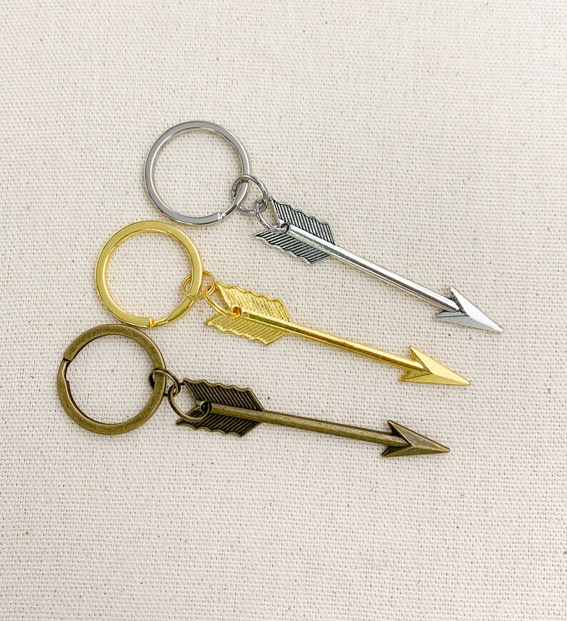 Op de afbeelding: Drie sleutelhangers met pijlpuntbedels. De sleutelhangers zijn zilver, goud en brons. De pijlpunten wijzen allemaal naar rechts.