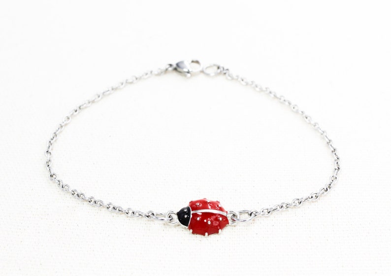 Puede incluir: Un brazalete de cadena de plata con un dije de mariquita roja. La mariquita tiene manchas negras y una cabeza negra.