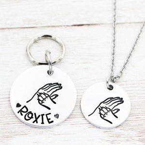 May include: A silver necklace and keyring set with a round pendant featuring a handprint design and the name "Roxie".