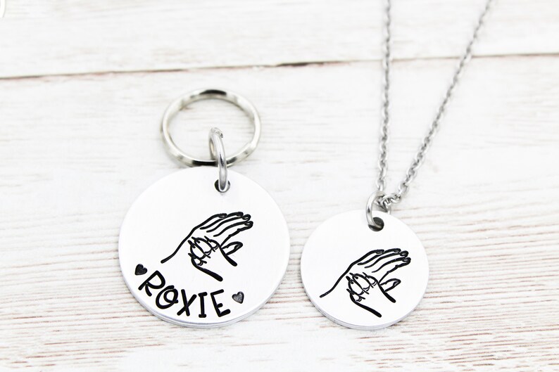 May include: A silver necklace and keyring set with a round pendant featuring a handprint design and the name "Roxie".