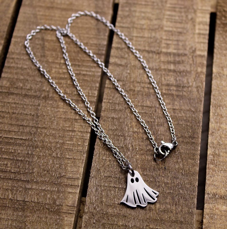 May include: A silver-colored necklace with a ghost pendant. The ghost pendant has two black eye holes and a flowing, sheet-like body. The chain is a delicate, linked design. The necklace is displayed on a wooden surface.